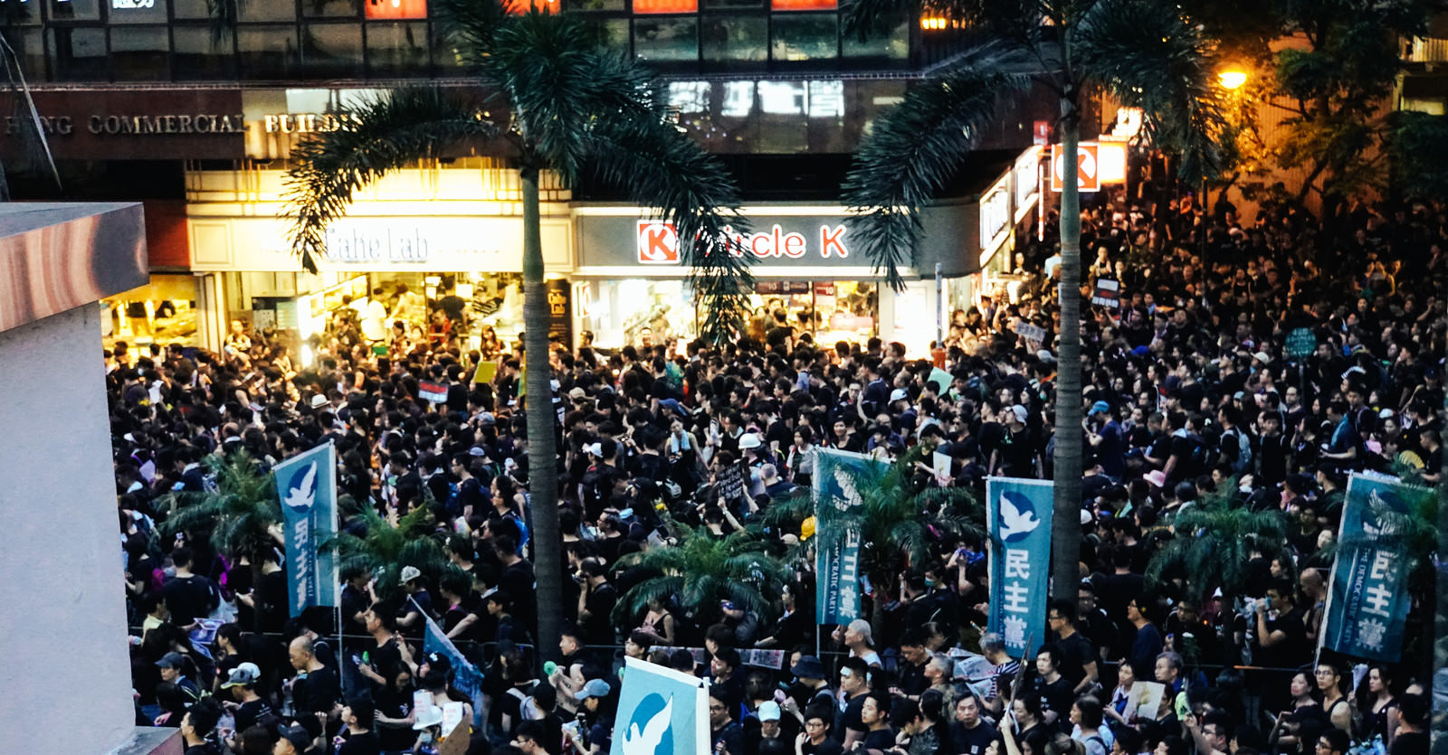 夜の香港の街頭で数千人規模の人群が密集し、青い横断幕を掲げて抗議活動を行っている光景