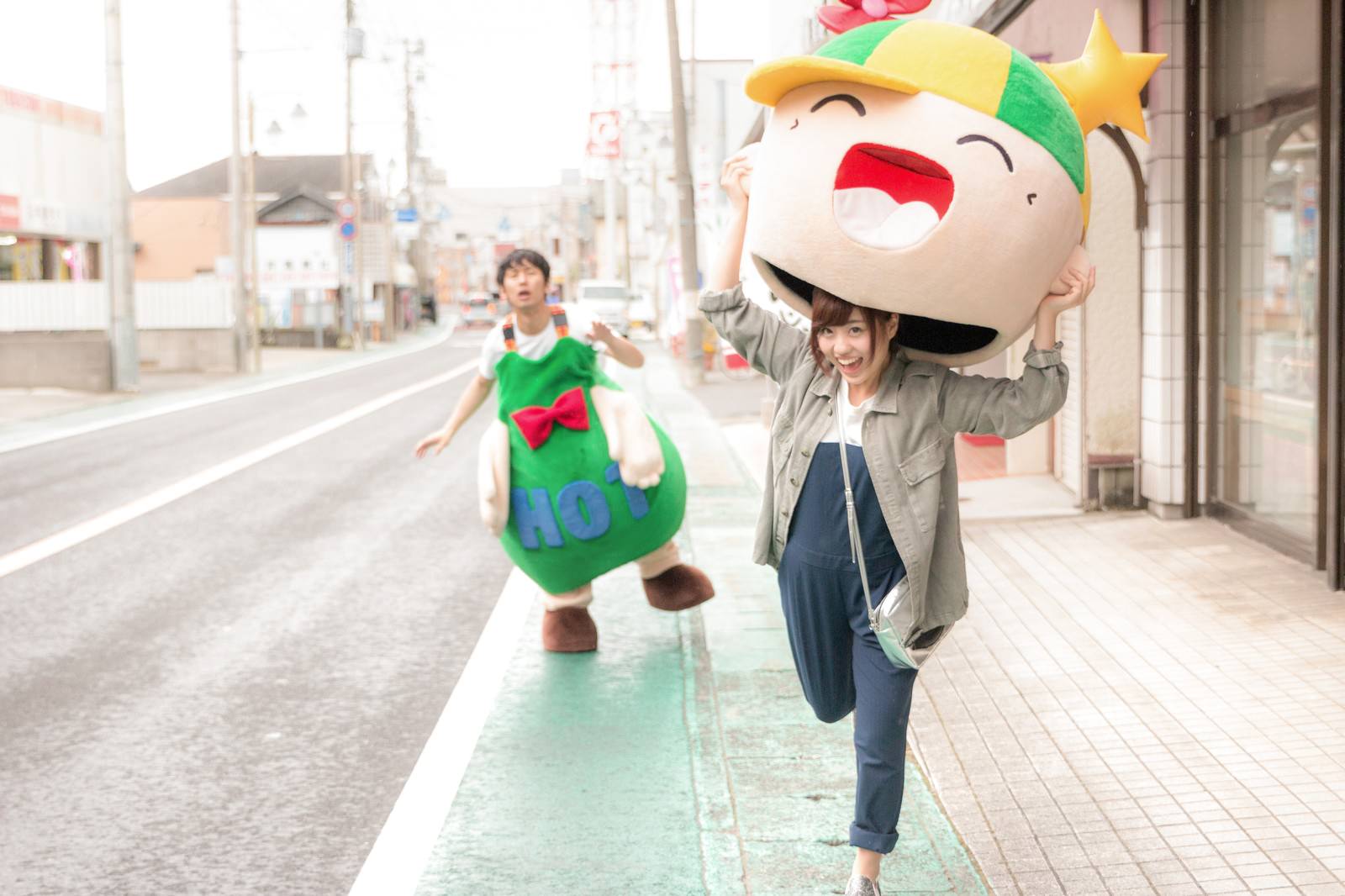 緑のきぐるみを着たゆるキャラの頭部を両手で持ち上げた女性と、後ろで驚く男性［モデル：ホットくん 大川竜弥 河村友歌］
