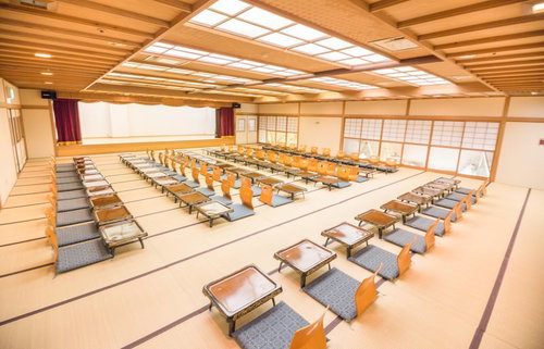 畳にオレンジの座布団と低いテーブルにお盆と湯呑が並ぶ大宴会場