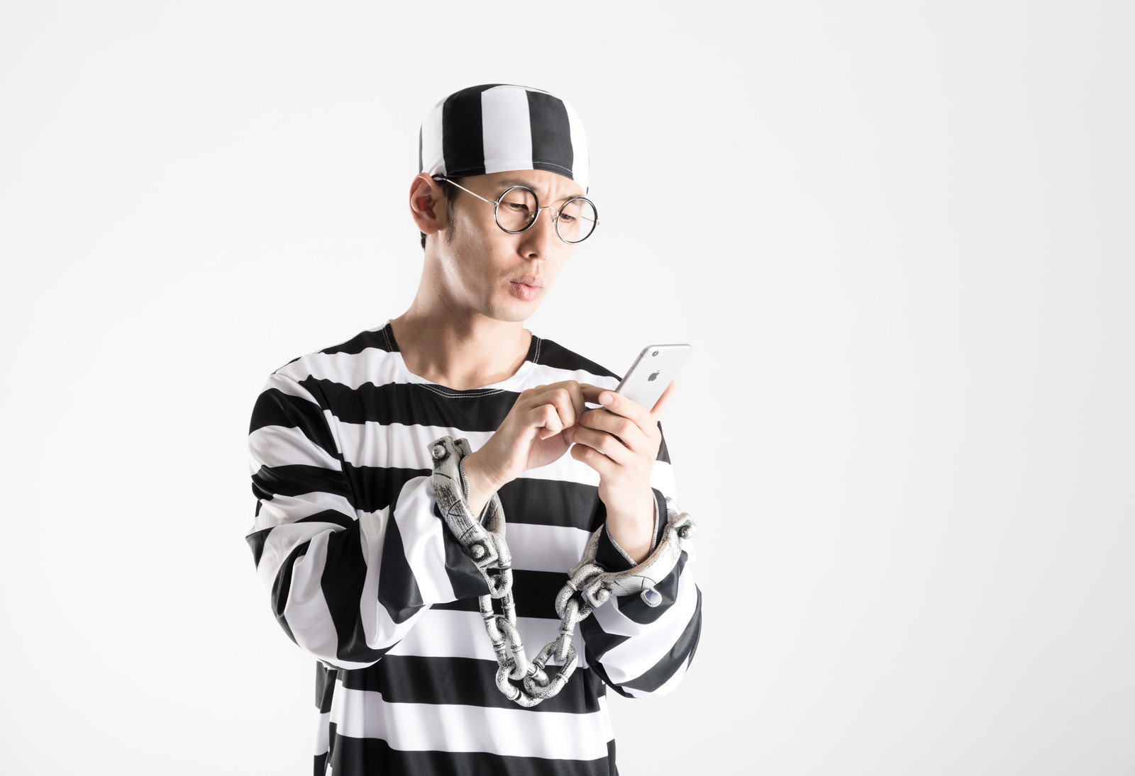 スマートフォンを操作する手錠をした囚人姿の男性。丸眼鏡と黒白縞服で横向きに下を向く［モデル：大川竜弥］
