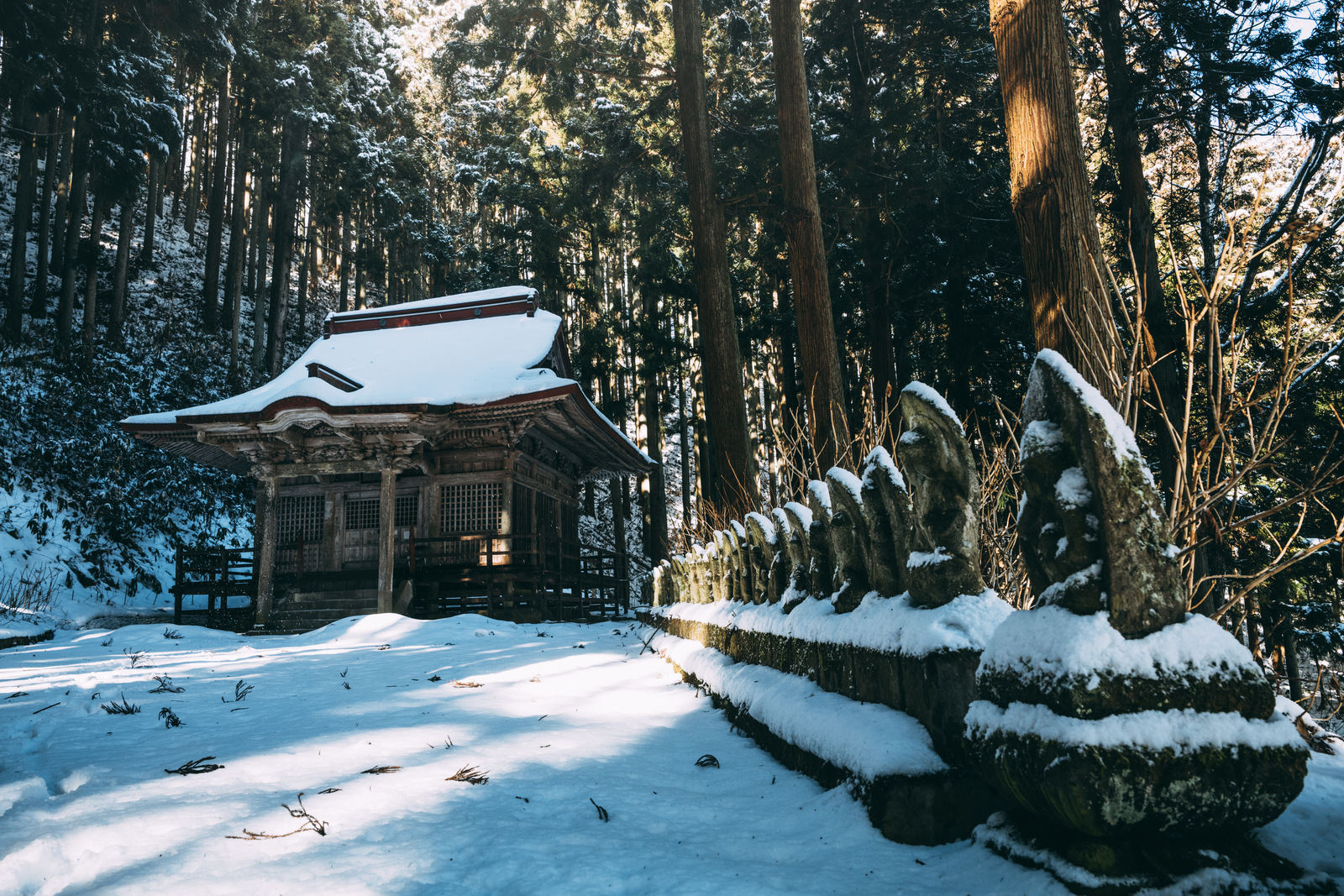 雪の積もった杉林の中に建つ観音堂と、並ぶ地蔵石像の冬景色