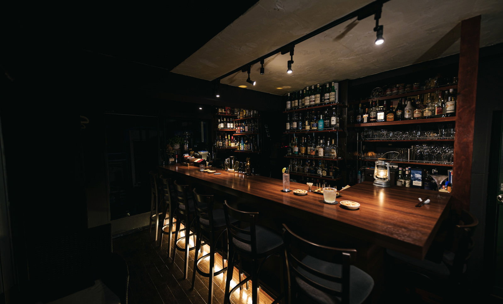 暗い照明の中、木製カウンターに椅子が並ぶバーの店内