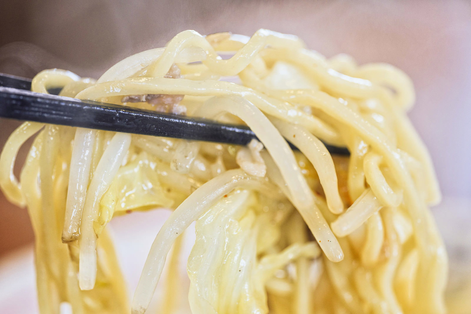 箸で持ち上げられた黄色いラーメンの麺から湯気が立ち上がっている