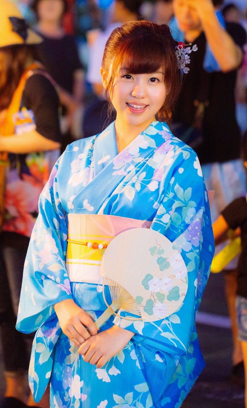 青い浴衣の女性が白いうちわを持ち笑顔で祭り会場に立つ［モデル：河村友歌］