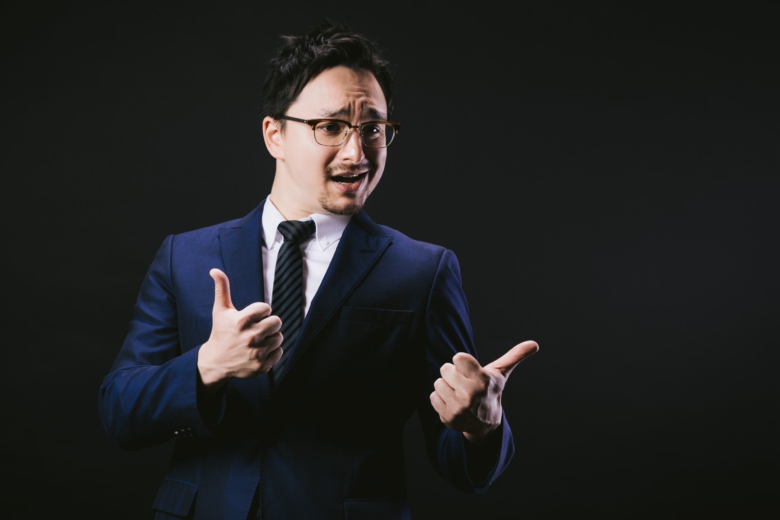 眼鏡をかけたスーツ姿の男性が興奮した表情で両手を上げ、両方の親指を立てるサムズアップポーズ［モデル：Max_Ezaki］