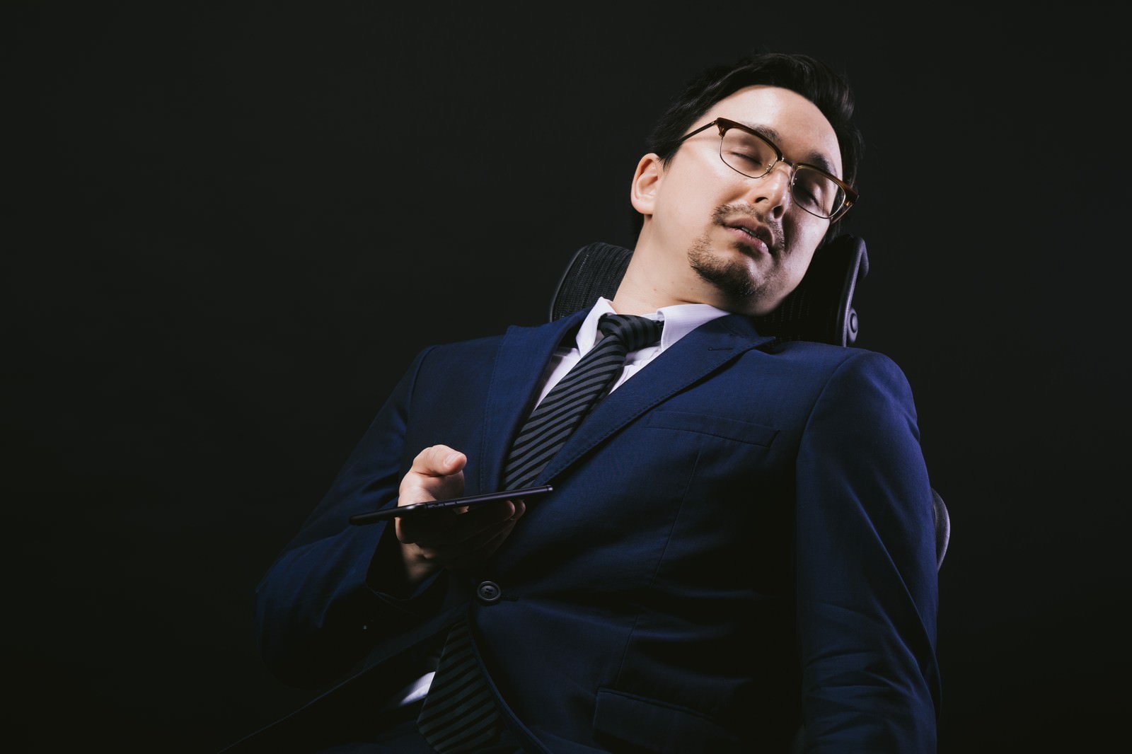 リクライニングチェアでスマホを持ちながら寝落ちするスーツ姿のドイツ人ハーフ男性［モデル：Max_Ezaki］