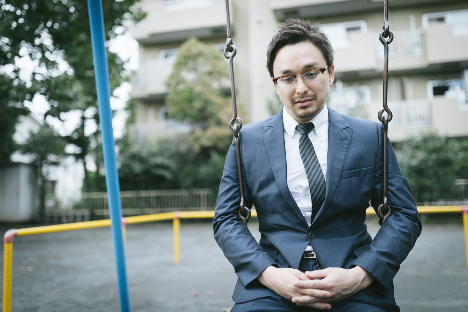 公園のブランコに座り憂鬱な表情を浮かべるスーツ姿の男性［モデル：Max_Ezaki］