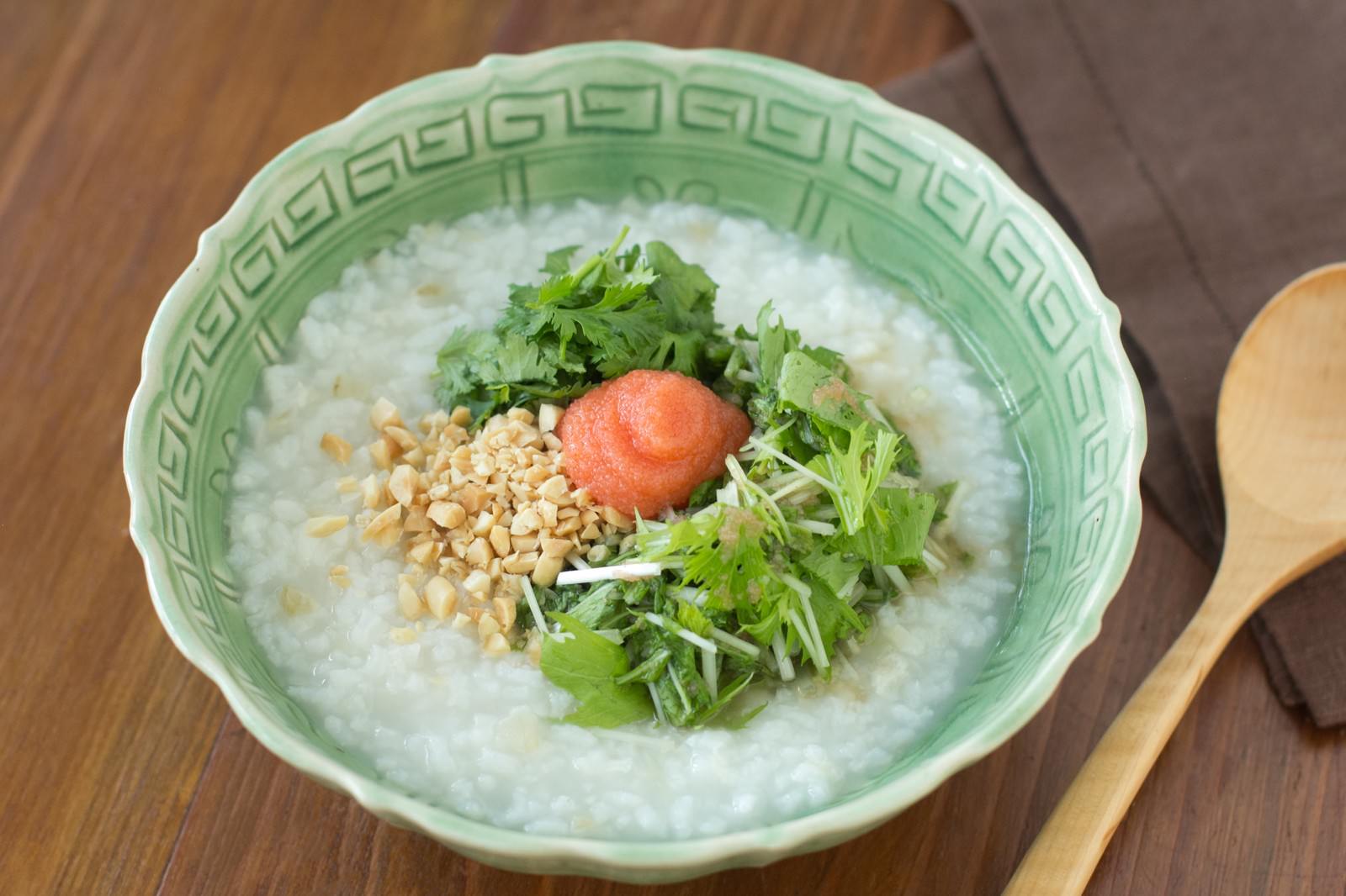淡緑色の磁器鉢に盛られた白いおかゆに、緑の香菜と水菜、赤い明太子、黄色いピーナッツがのっている香港がゆ
