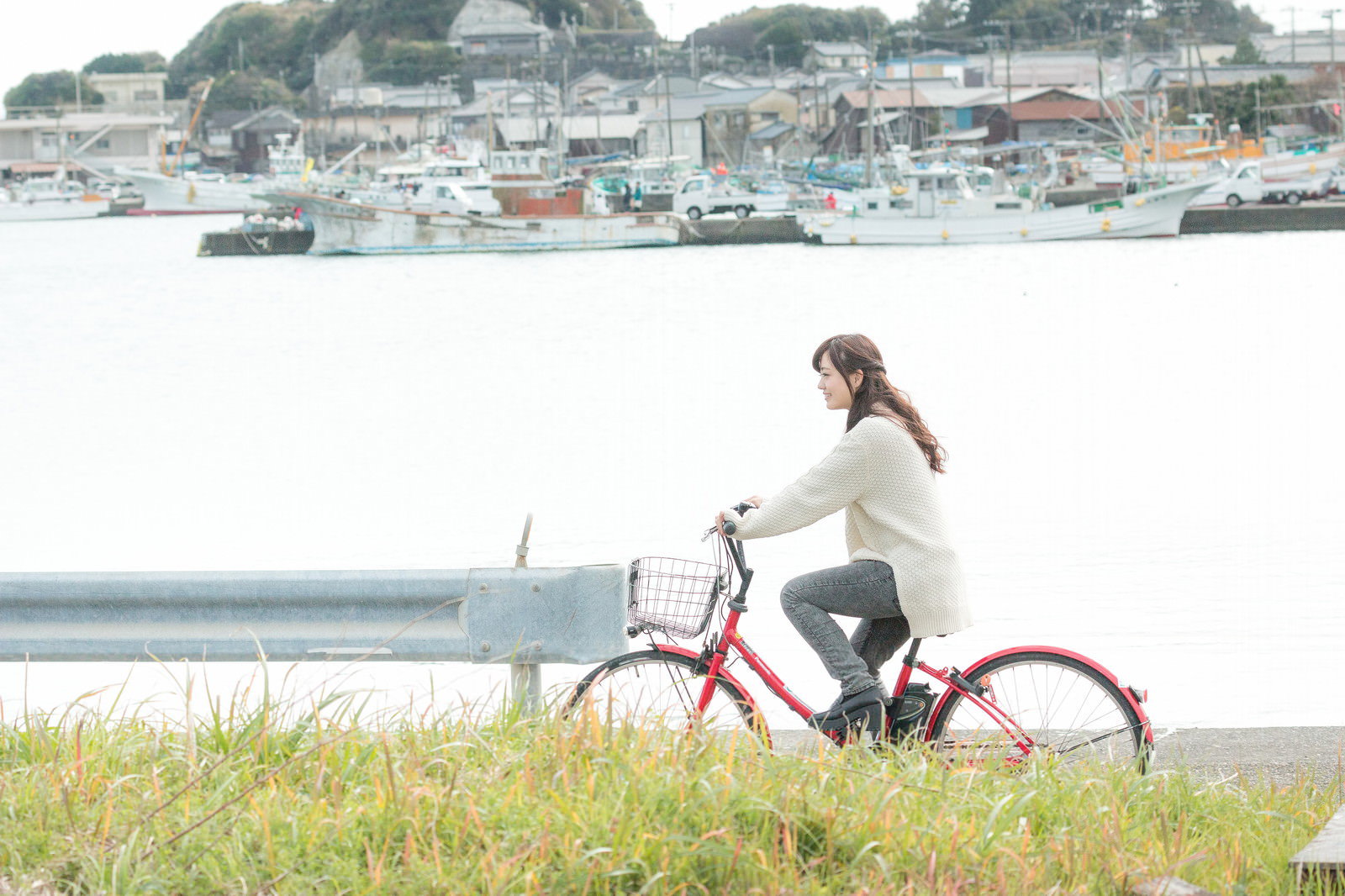 漁港を背景に赤い自転車に乗る白いニット素材の上着を着た女性［モデル：河村友歌］