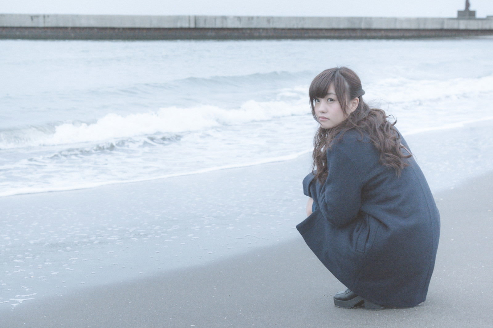 波打ち際の砂浜でしゃがみ込み、ふてくされた表情を見せる女性［モデル：河村友歌］