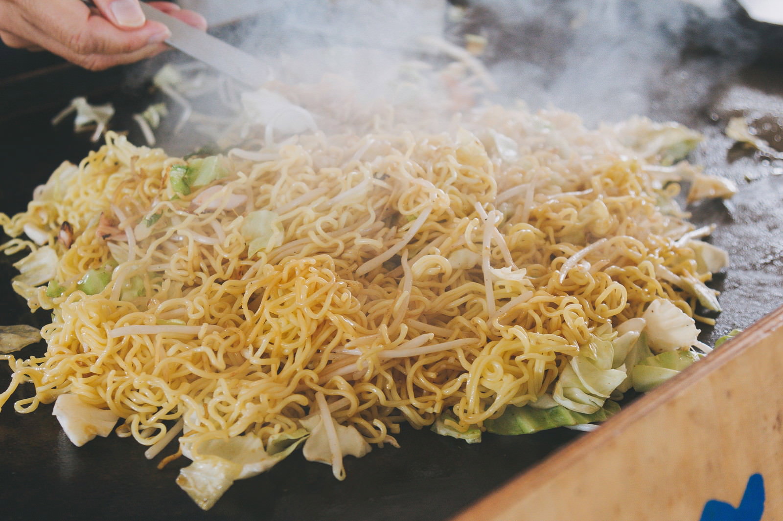 屋台の鉄板で調理された焼きそば。ねぎともやしが混ざり、トングで盛り付けられている