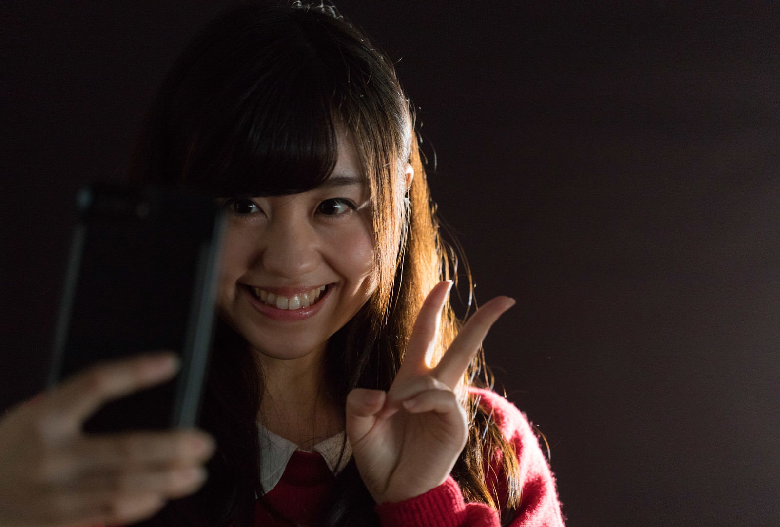 暗い場所でピースサインをしながら笑顔で自撮りをする女性［モデル：河村友歌］