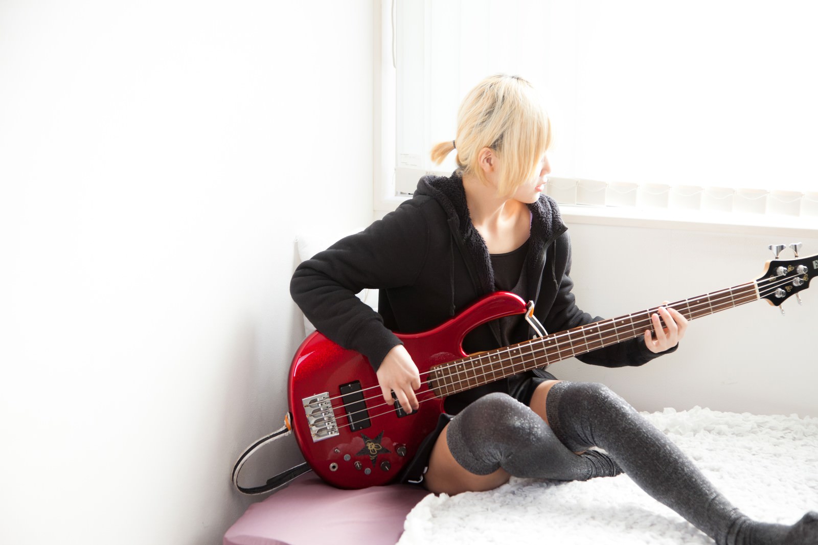赤いベースを演奏する女性がベッドの上で楽器を練習する様子［モデル：IRISMONDE］