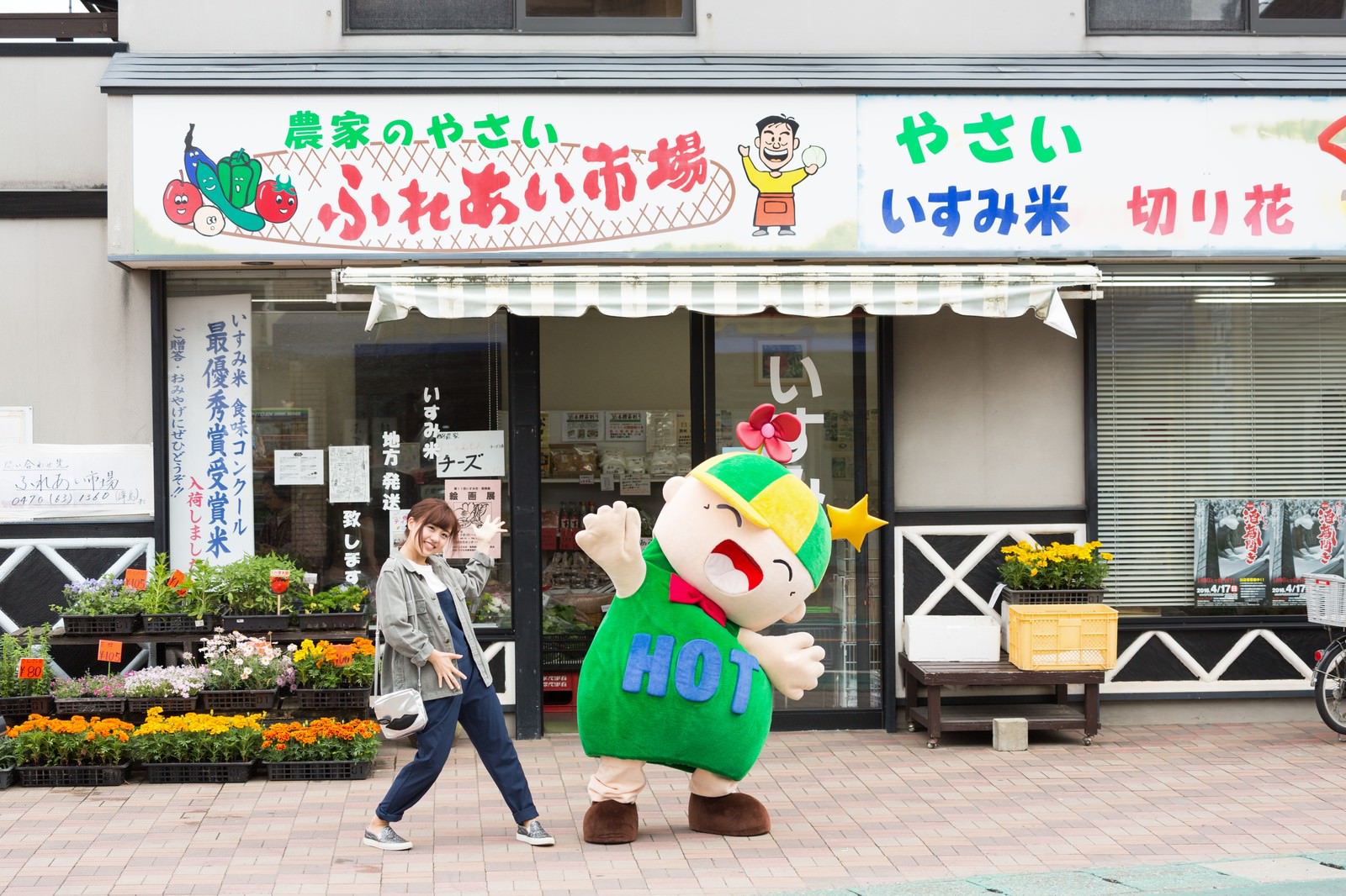 ふれあい市場の前に立つ女性と緑のゆるキャラ［モデル：ホットくん 河村友歌］