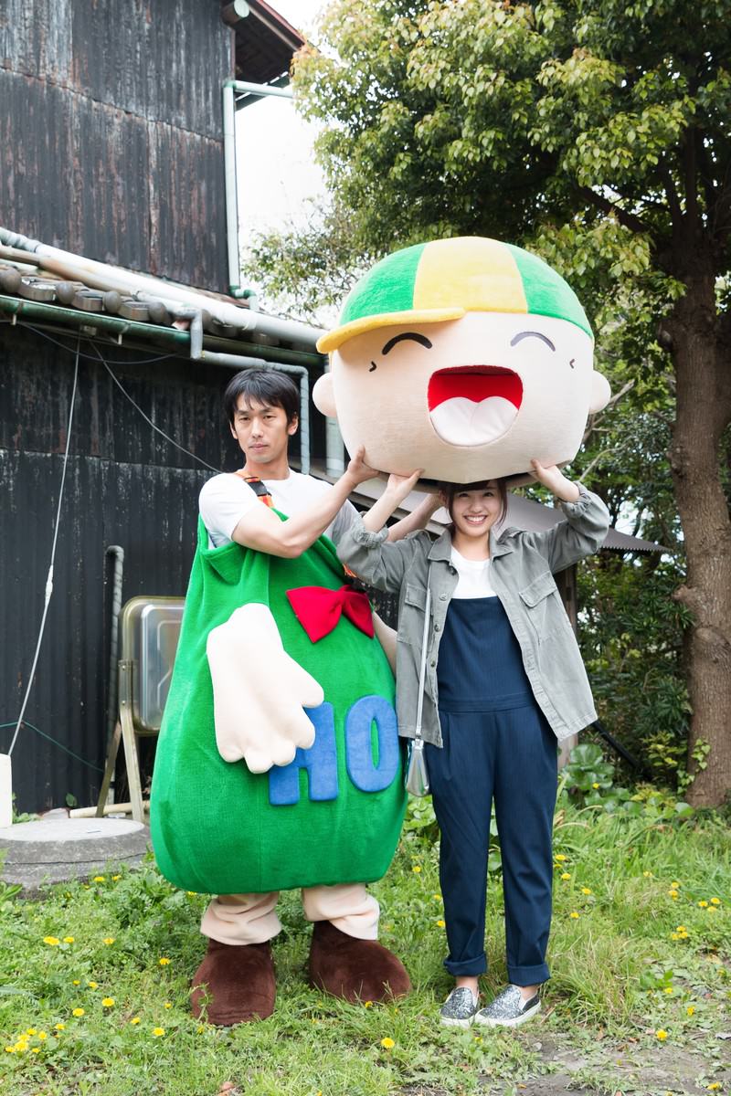 古い建物と樹木に囲まれた屋外で、緑色のゆるキャラ・ホットくんを背負う男性と女性が笑顔で肩を並べている［モデル：大川竜弥 ホットくん 河村友歌］