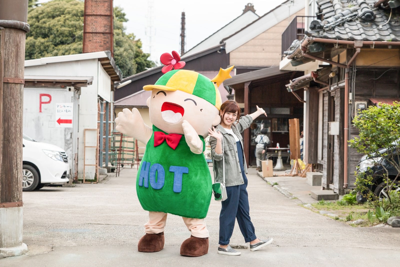 緑のマスコットキャラクターが両腕を広げて迎え、隣の女性が手を上げている大原中央商店街の風景［モデル：ホットくん 河村友歌］