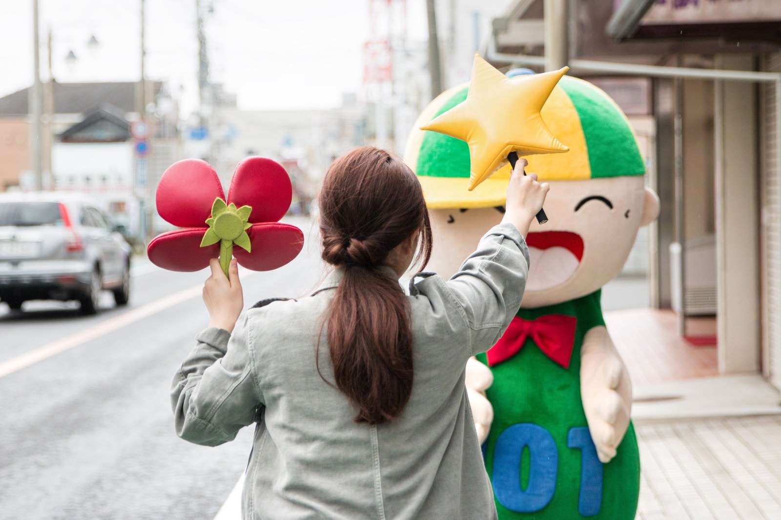 商店街でゆるキャラに赤い花のおもちゃを見せる灰色ジャケットの女性［モデル：ホットくん 河村友歌］