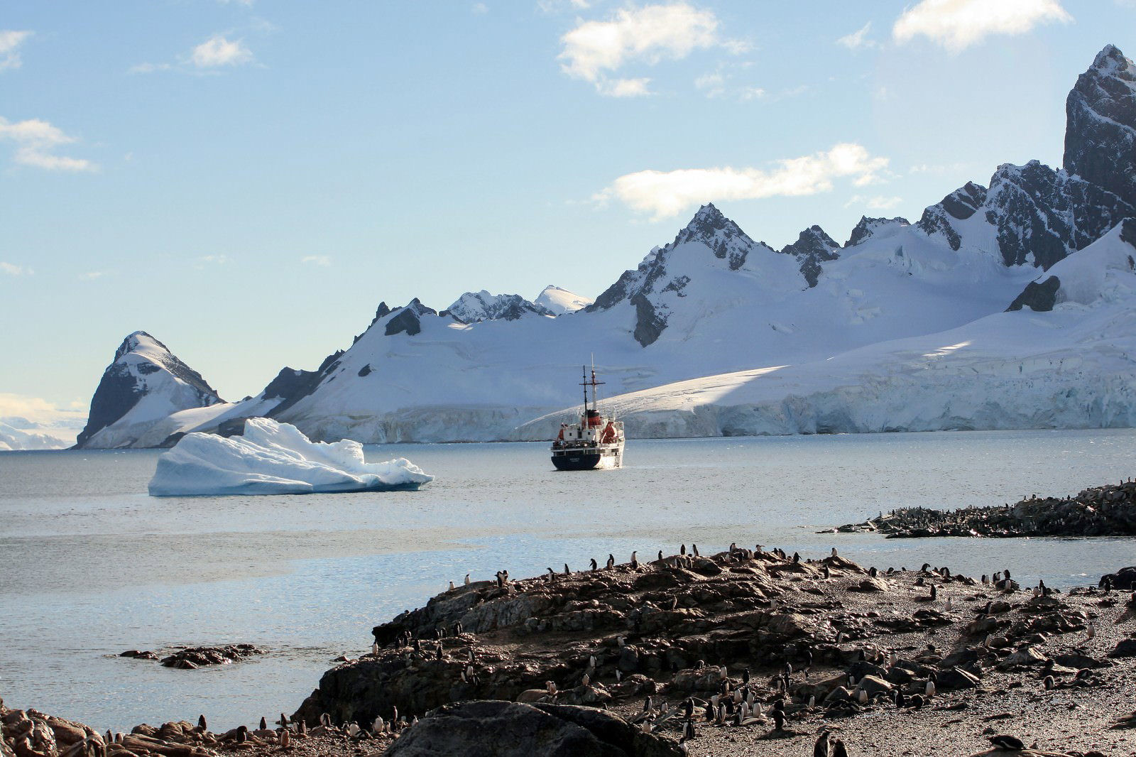 雪をかぶった険しい岩峰と流氷、ペンギン群、観測船が見える南極の風景
