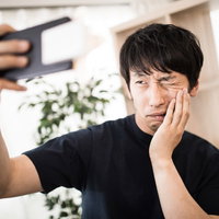 虫歯痛で頬を押さえながらスマホで自撮りする男性の写真