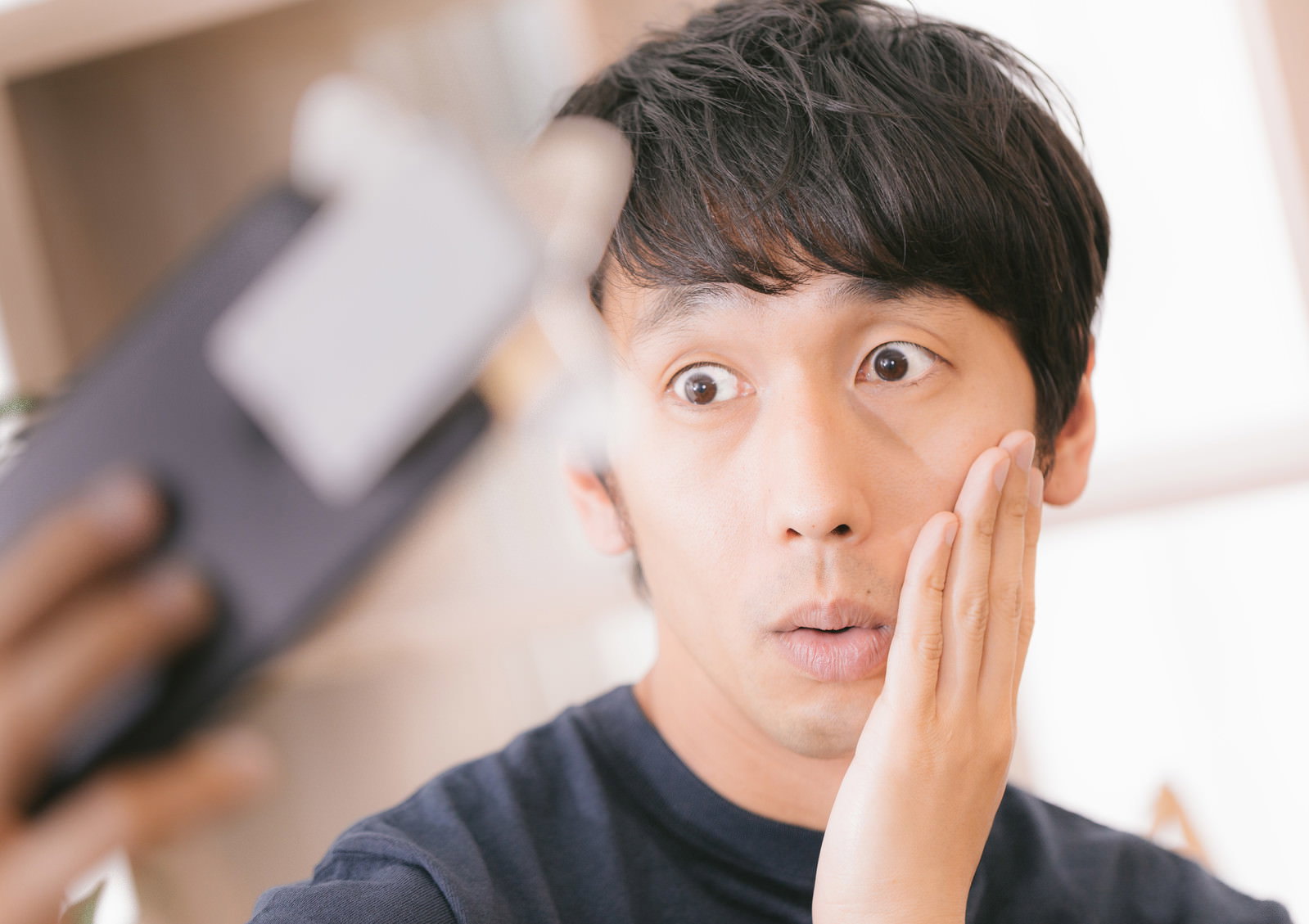 スマートフォンを持ちながら頬杖をついて驚いた表情を見せるアラサー男性［モデル：大川竜弥］