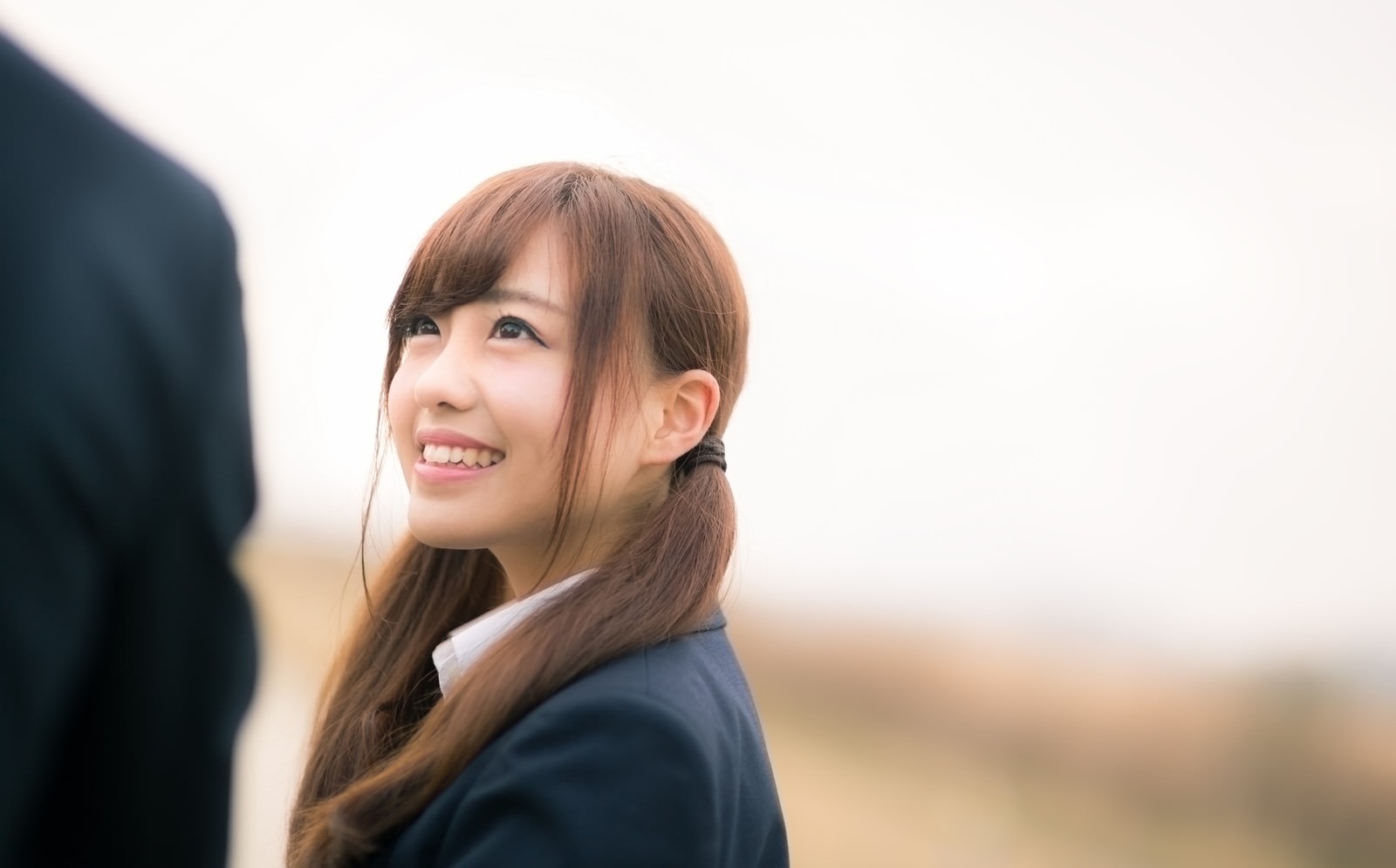 笑顔で上を見上げるツインテールの紺ブレザー制服の茶髪女性［モデル：河村友歌］