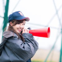 野球場のフェンス前で赤いメガホンを持ち笑顔のグレージャケットの女性の写真