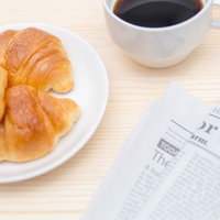 クロワッサンとコーヒーカップと英文の新聞が並ぶ朝食風景の写真