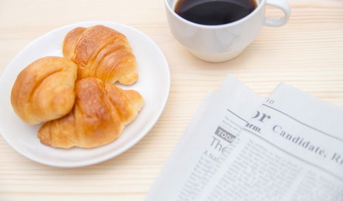 クロワッサンとコーヒーカップと英文の新聞が並ぶ朝食風景