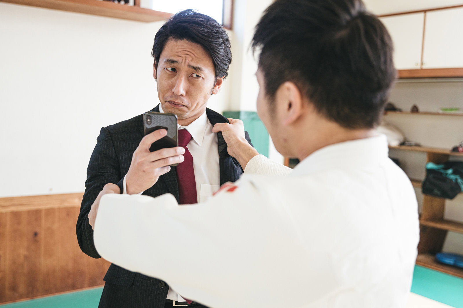 スマホ画面を見るスーツ姿の男性と、道場で組み手を取る柔道着姿の男性［モデル：大川竜弥］