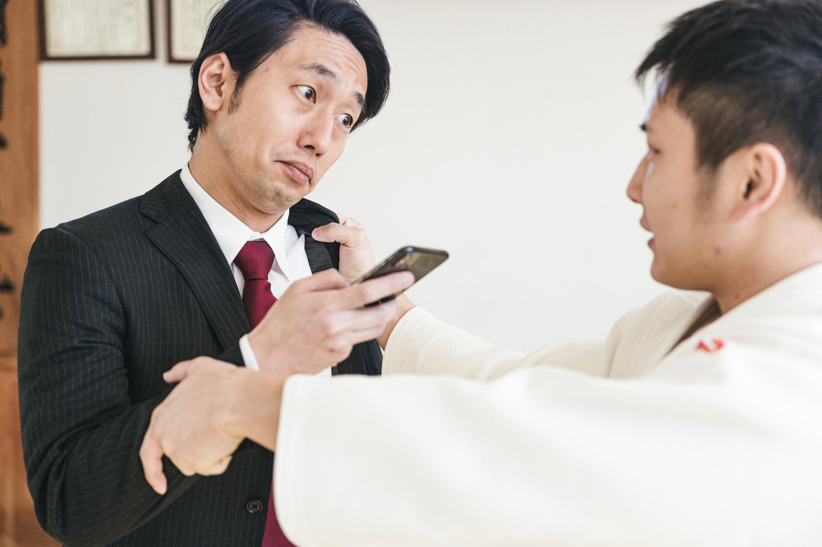 スマートフォンを持ち強がるスーツ姿の男性と道場で対峙する場面［モデル：大川竜弥］