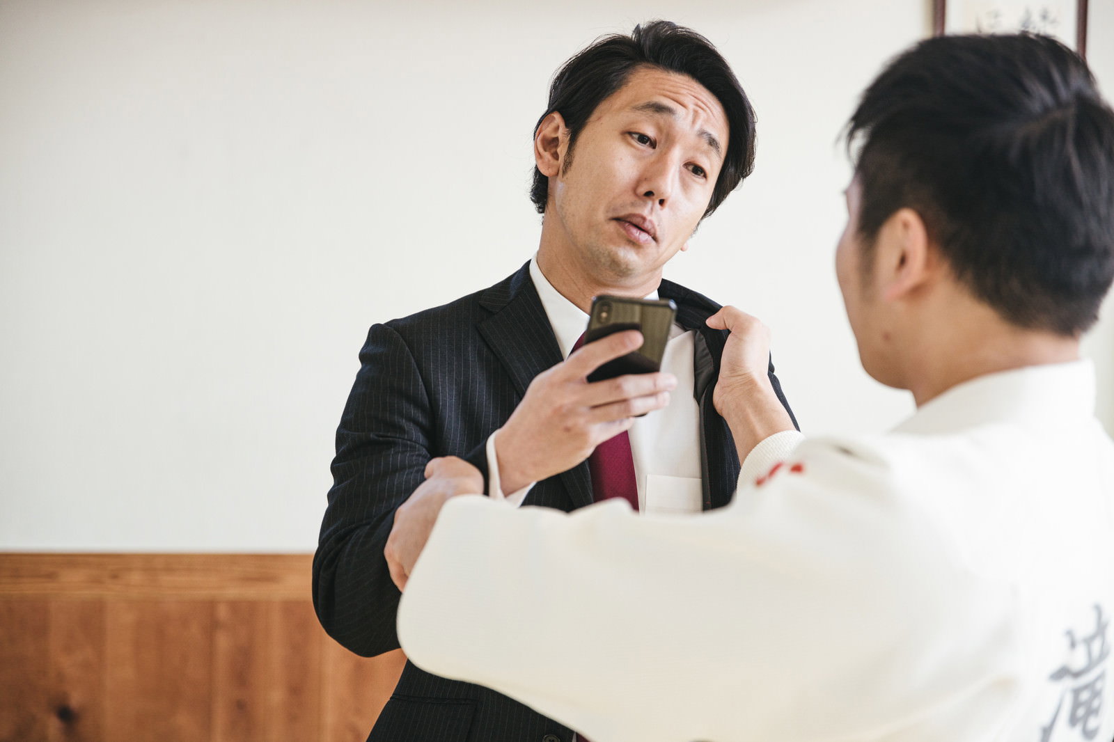 スマホを突きつけるスーツ男性と柔道着の人物［モデル：大川竜弥］
