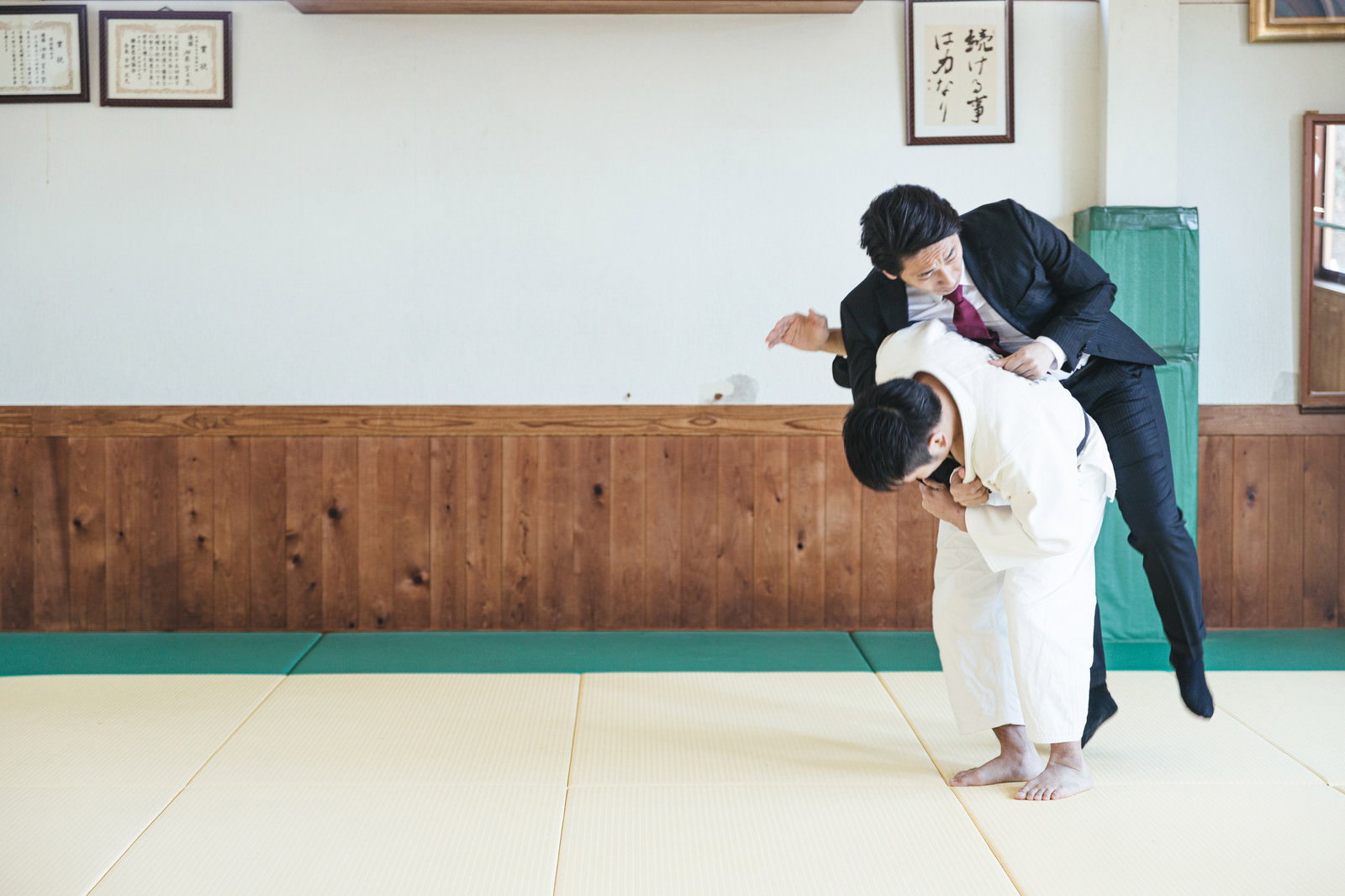 道場で柔道着の人物がスーツ姿のサラリーマンを一本背負いで投げている［モデル：大川竜弥］