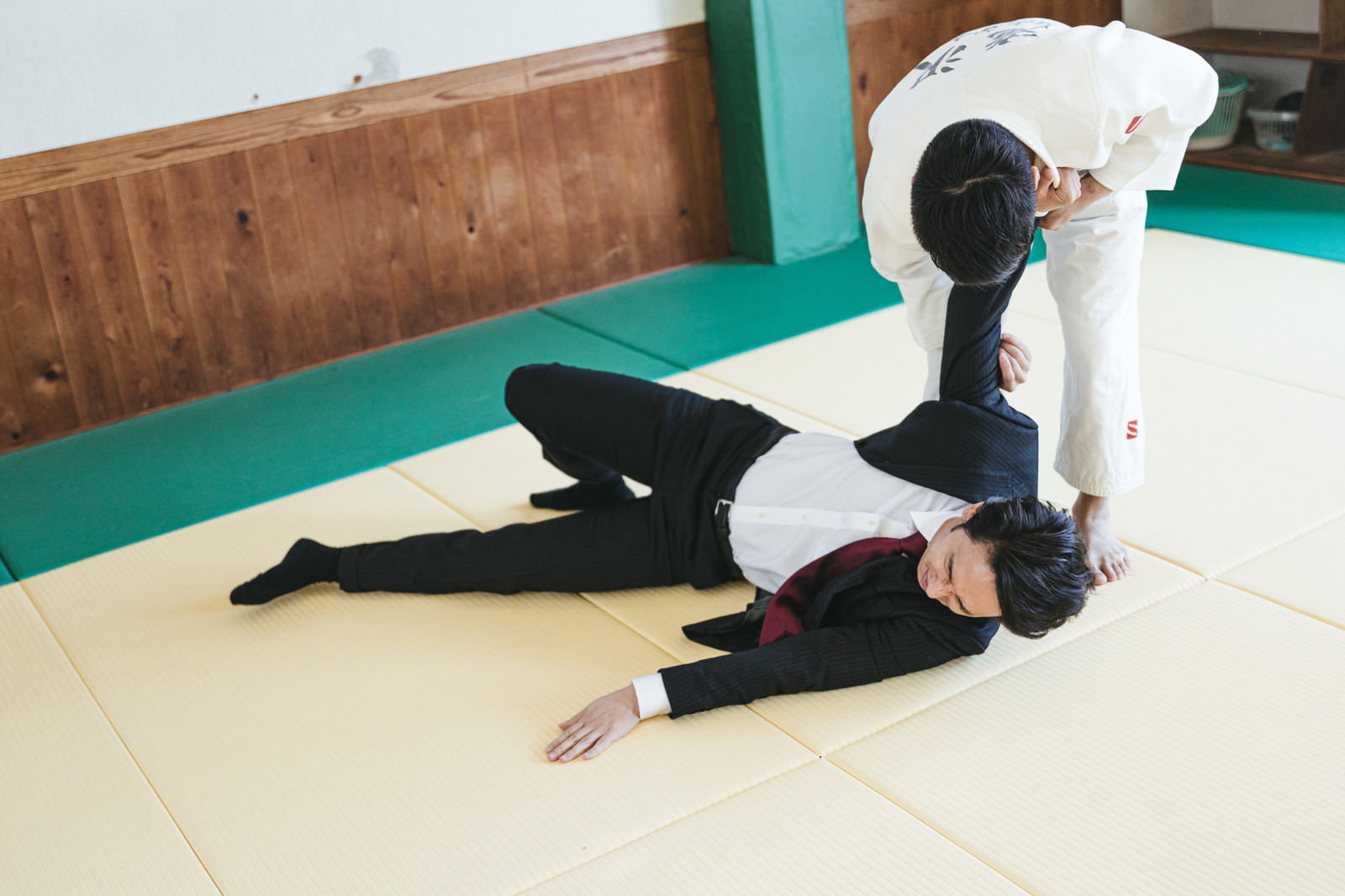 道場で柔道着の男性に一本背負いで投げられるスーツ姿の会社員