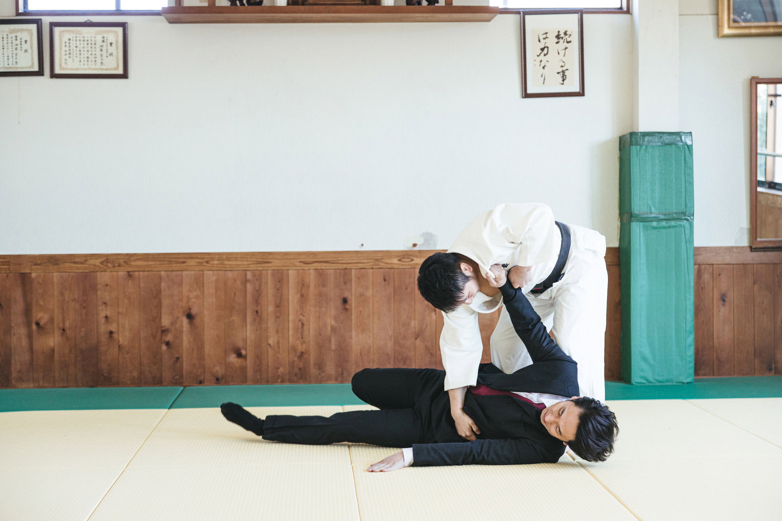柔道場の畳の上でスーツ姿のサラリーマンが柔道着の男性に投げ技をかけられている様子［モデル：大川竜弥］