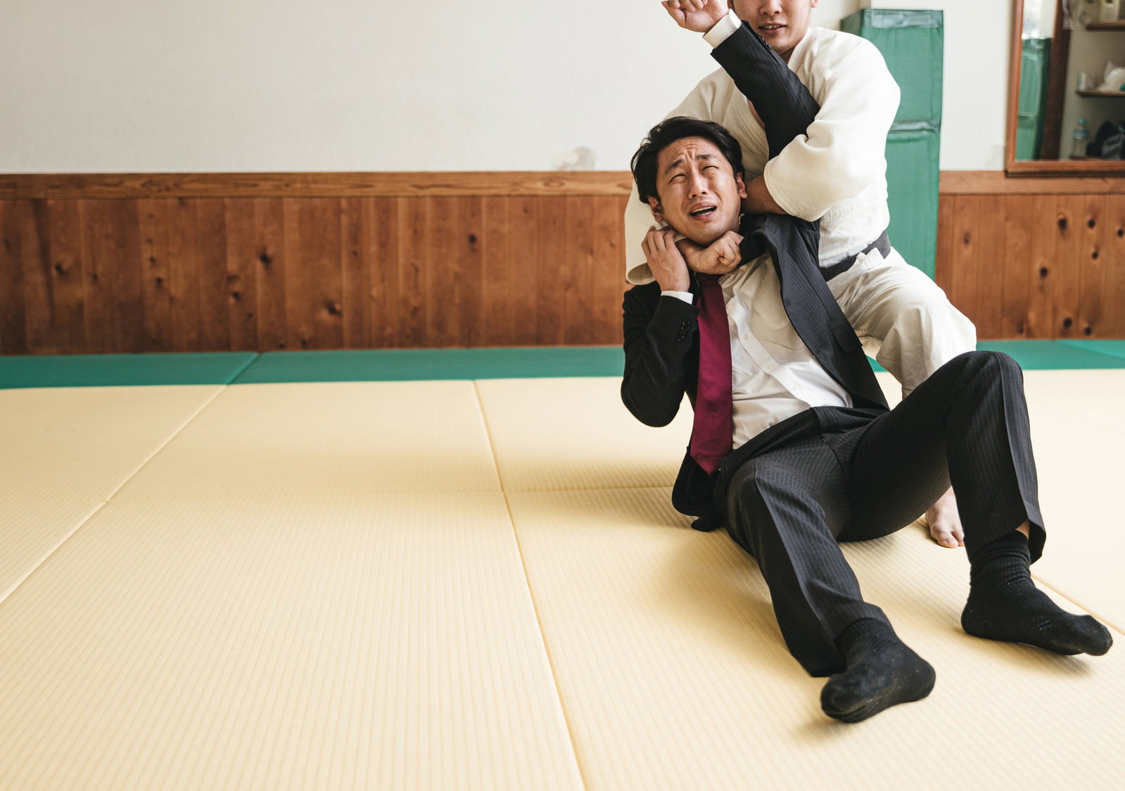 畳の上でスーツ姿の男性が片羽絞めから逃れようと苦しそうにもがいている様子［モデル：大川竜弥］