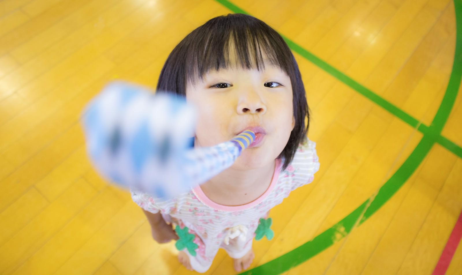 ピロピロを吹く子ども。黄色背景の前でピンク色の服を着た女の子がおもちゃを吹いて遊んでいる［モデル：あんじゅ］