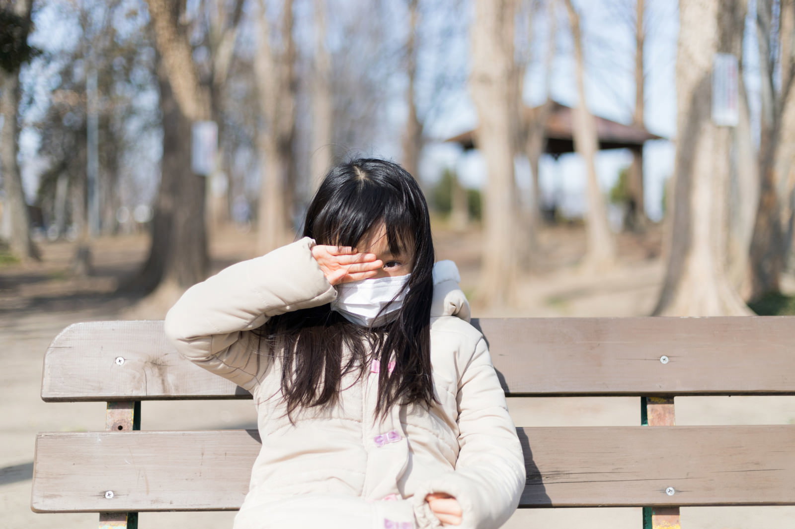マスクを着用した女性がベンチに座りながら目をこすっている様子［モデル：あんじゅ］