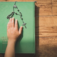 緑色の聖書に手を置き誓う子供の手とロザリオの写真