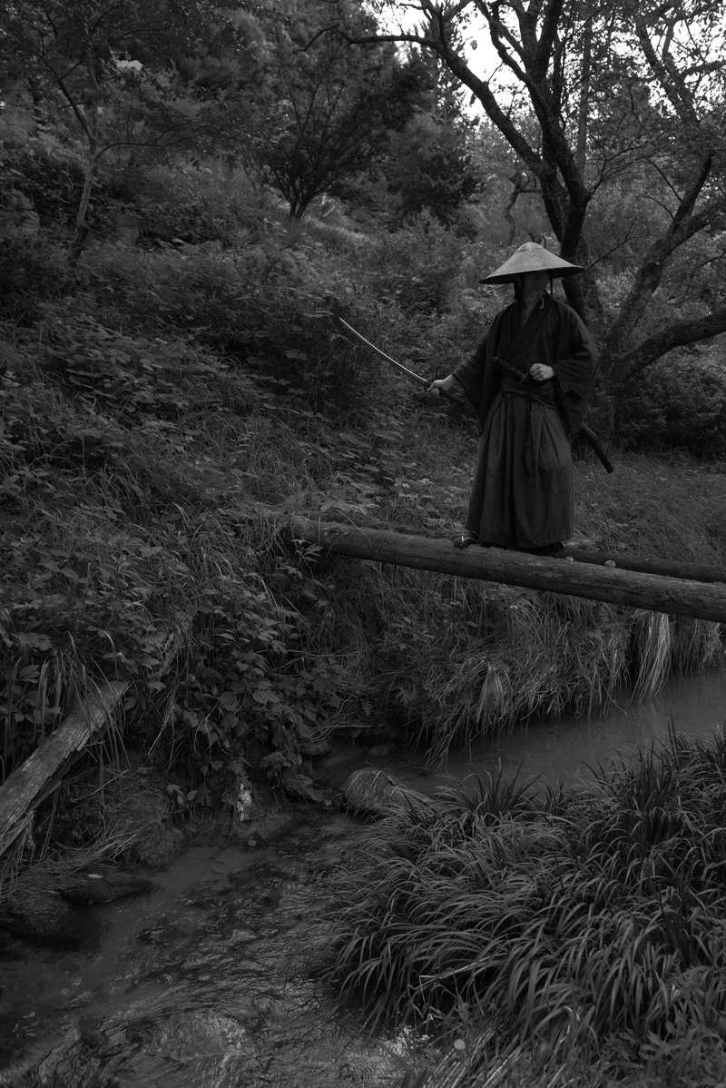 黒い着物と袴、笠を被り刀を構える侍が丸太橋に立つ［モデル：まーこ］