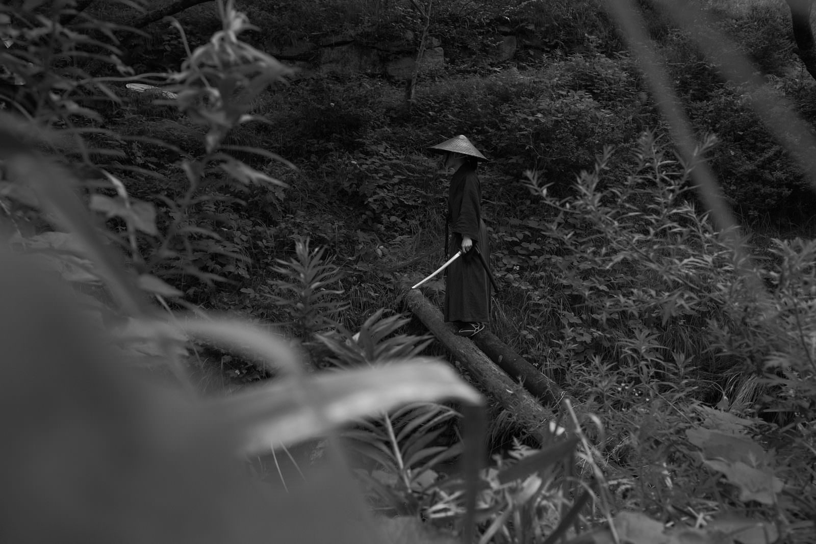 草陰から丸太橋を通る追手を待つ、刀を持った黒装束の男性［モデル：まーこ］