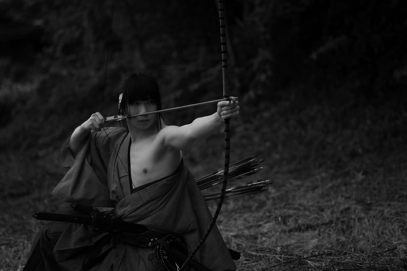袴姿の男性が森の中で大弓を引いている様子を撮影したモノクロ写真［モデル：まーこ］