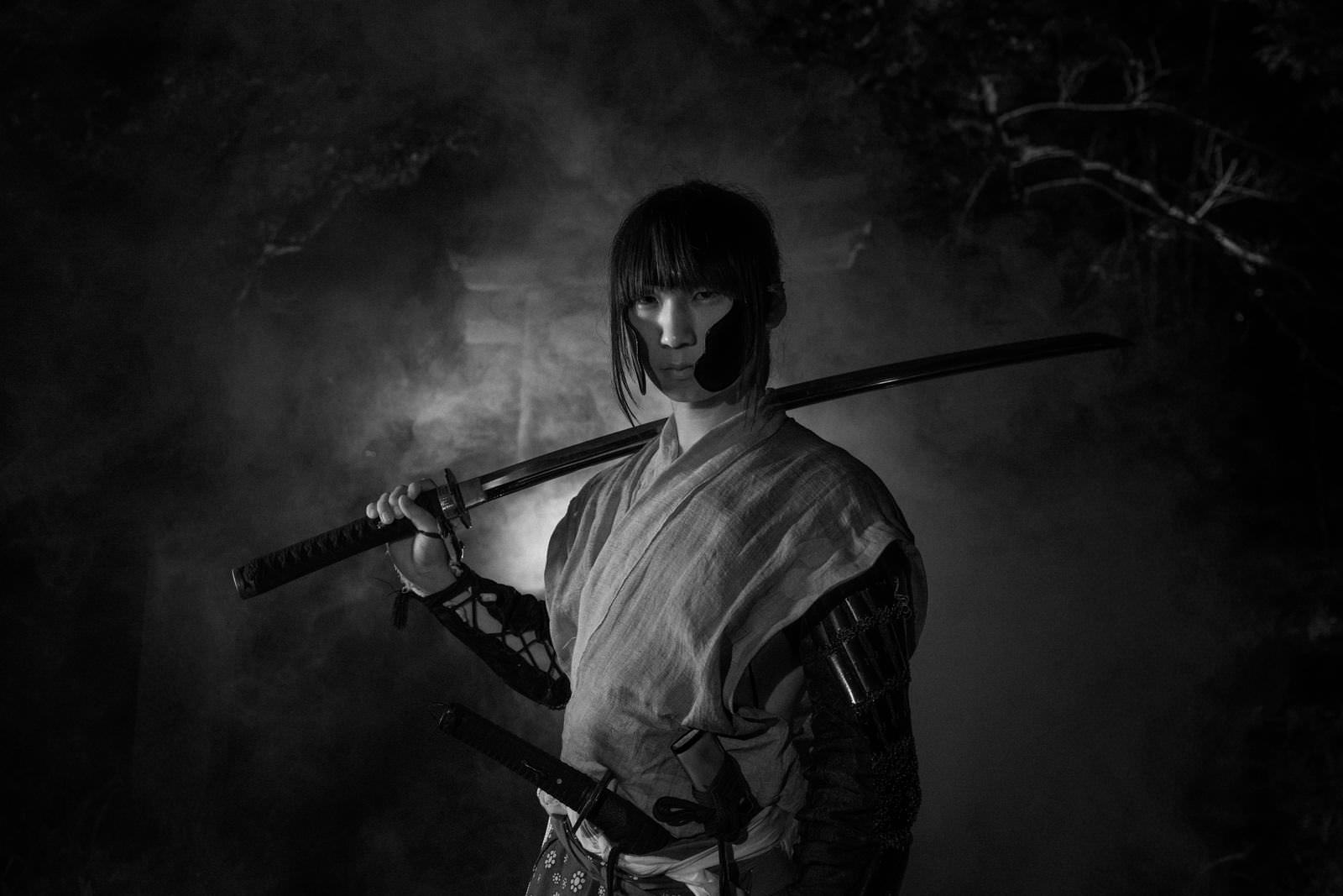 煙の中で刀を肩に担ぐ武士の男性が横向きで立つモノクロ写真［モデル：まーこ］