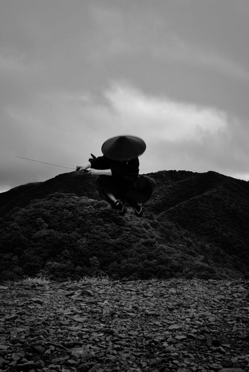 袴を着た侍が岩の上から空中にジャンプしているモノクロ写真［モデル：まーこ］