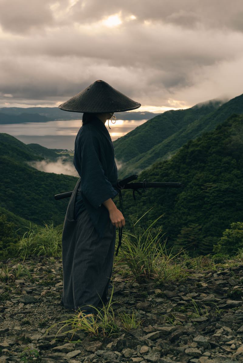 御霊櫃峠で厚い雲に覆われた夕空を見つめる袴と笠を身に着けた侍［モデル：まーこ］