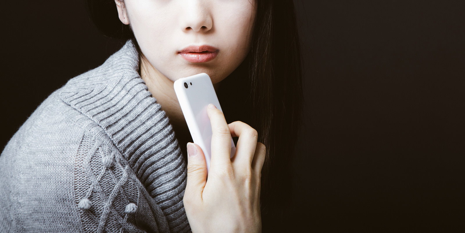 グレーのニットを着た女性がスマートフォンを顎に当てて考えている様子