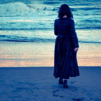 夕暮れの波打ち際を歩く、寂しげな女性の写真