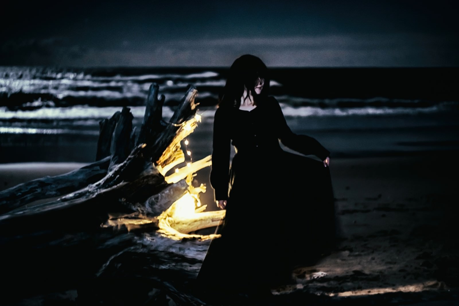 夜の砂浜で流木の炎の前に立つ黒い服の女性