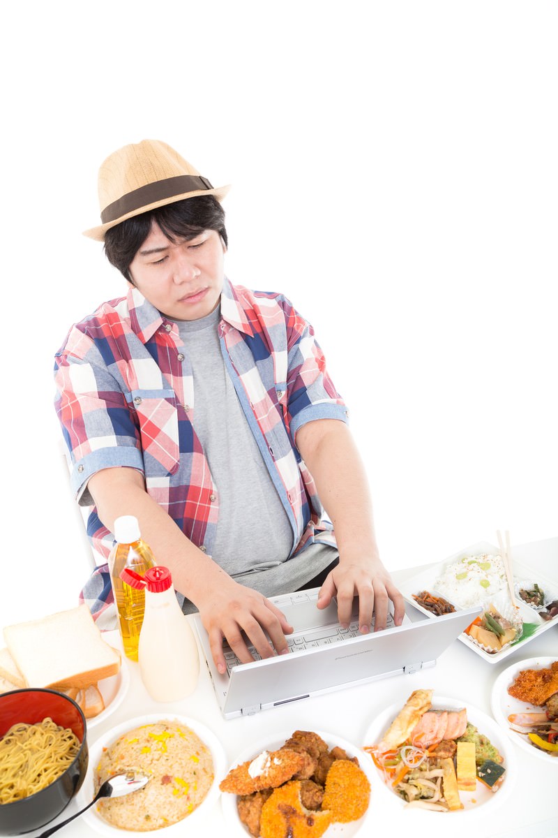 ノートパソコンの前に並ぶ出前の食事をながら食べする男性［モデル：あまのじゃく］