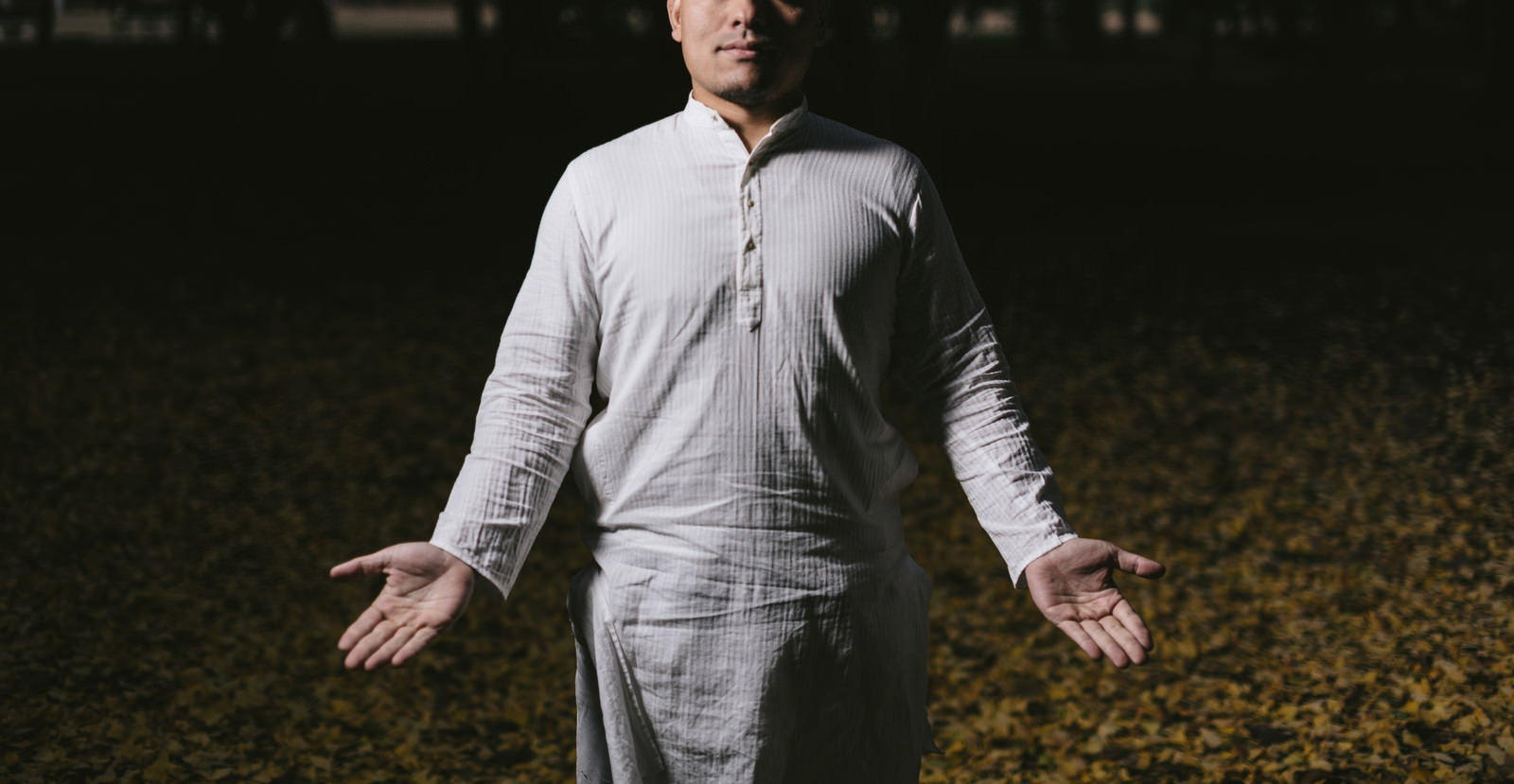 白いシャツの男性が両腕を広げ、目を閉じた状態で立つ。背景は暗く、人物に光が当たっている