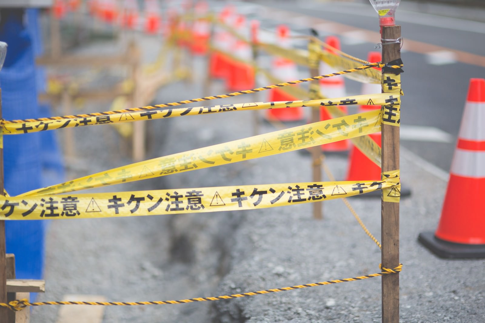 道路工事現場に張られた黄色と黒の注意テープが木の支柱に巻き付けられ、背景にカラーコーンと進入禁止フェンスが配置されている
